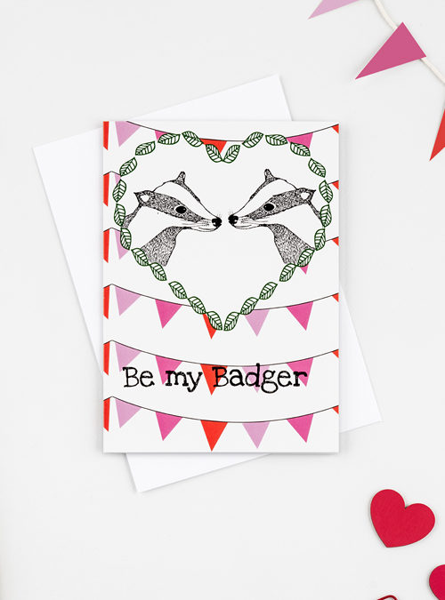 Photograph of a Badger card. Illustration of 2 badgers facing each other surrounded by a leaf heart with pink and red bunting in the bacjground. The wording 'Be my badger' in black ink underneath