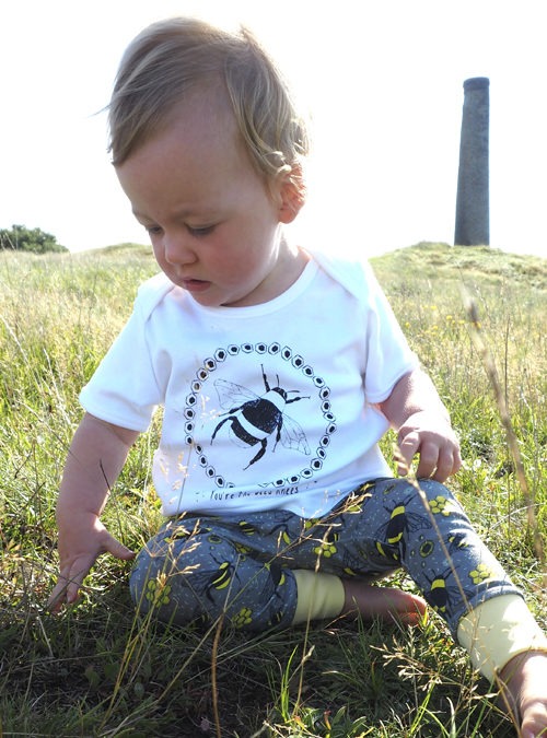 Baby model wearing Bee outfit. White T-shirt featuring a black print of a bee paired with grey leggings featuring a repeat pattern of bees, honeycomb and flowers. Baby sitting in the grass up and looking down