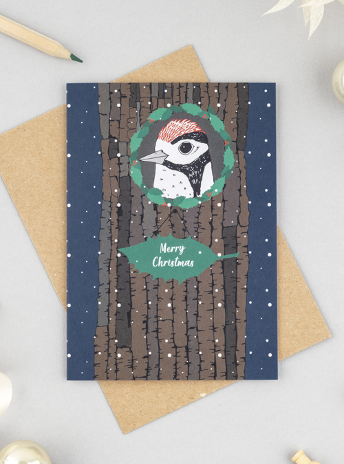 Photograph of a Christmas card with a woodpecker on the front in his home with a holly wreath around the hole and a big piece of holly saying 'Merry Christmas'